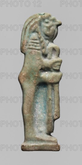 Amulet — Relief figurine of Kebehsenuf, one of the Sons of Horus. Kebehsenuf is depicted in right profile with a mummy-like silhouette and a falcon-like head covered with a wig, holding a scroll of fabric in front of him. The back of the figurine is smooth with two holes for attachment. This object forms a set of four Sons of Horus with inventory numbers 238258–238260. Late Period–Ptolemaic Period, 664–30 BC, Egypt. Amulet; height 4.6 cm, width 1.4 cm., A small carved figurine viewed in profile showing a standing humanoid figure with a stylized head and elongated headdress, holding a smaller rounded figure against its chest; the surface shows a weathered, slightly rough texture with patches of worn areas and pitting, and the visible colors include shades of green, turquoise, pale blue, tan, beige, and hints of brown against a plain light gray background.