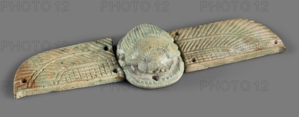 Winged scarab, Winged scarab with a realistically modeled torso, head, and legs. On the outstretched wings, imitating plumage, is an ornament consisting of a grid-covered field and two stripes covered with diagonal lines. The small oval base on which the scarab is placed has six holes for attachment to a mummy net. There are three similar holes on each wing. Due to the non-corresponding holes on the sides of the scarab and on the inner part of the wings, it can be assumed that they were not originally a single piece, but were fitted together later for antiquarian purposes. 664–332 BC, Late Period, Egypt. Amulet scarab. Height 3.4 cm, width 12.5 cm., A carved object composed of a central rounded piece flanked by two elongated wing-like pieces, all resting on a flat gray background; the central piece has a textured, ridged surface and a rounded protruding form with carved indentations and holes, showing a weathered surface with shades of pale green, turquoise, beige, and areas of brown; ...