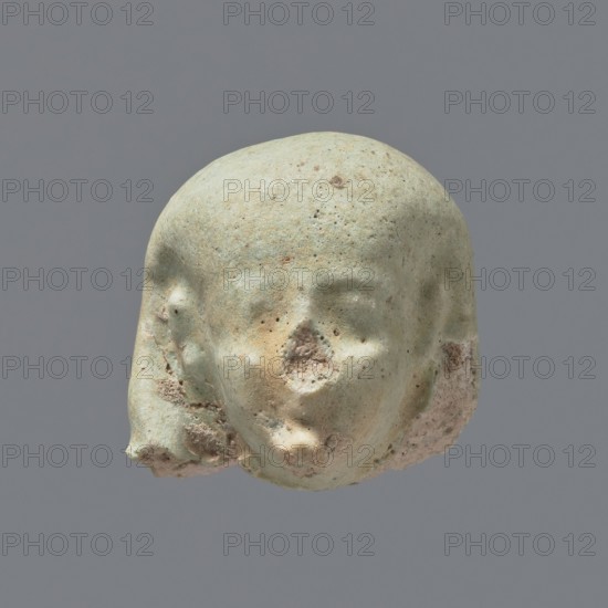 Fragment of a shabti. Head of a shabti figurine with not very carefully rendered facial features, in a smooth wig and with a thin beard not connected to the neck. Celadon glaze. 332–30 BC; Ptolemaic Period. El-Kantara. Shabti, height 2.5 cm., A small sculpted head fragment with a rounded top and stylized facial features, rendered in a pale greenish hue with areas of beige and brown discoloration; the surface shows pitting and rough texture, especially around the nose and lower jaw where some erosion has exposed a sandy, pinkish-beige interior; the left and right sides of the head have irregular chipped areas with a mix of green, beige, and pinkish tones; the background is a uniform medium gray.