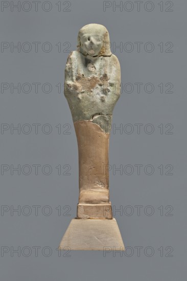 Fragment of a shabti, Mummy-shaped funerary figurine shabti, grave good, upper part preserved to the beginning of the thighs. On the head is a smooth tripartite wig; hands hold hoes; a back pillar is present. Facial features are carelessly rendered. Celadon glaze. Ptolemaic Period, 332–30 BC; from El-Kantara. Height 7.8 cm., A small sculpted figure with a rounded head and simplified facial features atop a tapered body mounted on a rectangular stepped base; the upper portion shows an eroded, rough surface with patches of pale green and light brown, while the lower shaft and base have smoother surfaces in beige and tan tones against a solid gray background.