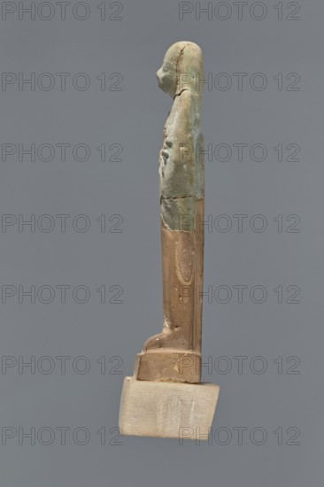 Fragment of a shabti. Mummy-shaped figurine, grave good, preserved to the beginning of the thighs. Upper part of a shabti figurine with a smooth tripartite wig, holding hoes, and a back pillar; facial features are carelessly rendered. Celadon glaze. Ptolemaic Period, 332–30 BC; from El-Kantara. Shabti, height 7.8 cm., A slender vertical figurine viewed in profile against a solid gray background, with a rounded head and a tapered torso that shows surface irregularities and small dents; the upper portion of the figure has a greenish-blue patina with areas of lighter green and pale blue, while the lower portion is a warm brown with smoother texture and remnants of a lighter beige base at the bottom; overall the object shows seams and cracks where the colors meet and a subtle contrast between the cool green-blue and the warm brown and beige tones.