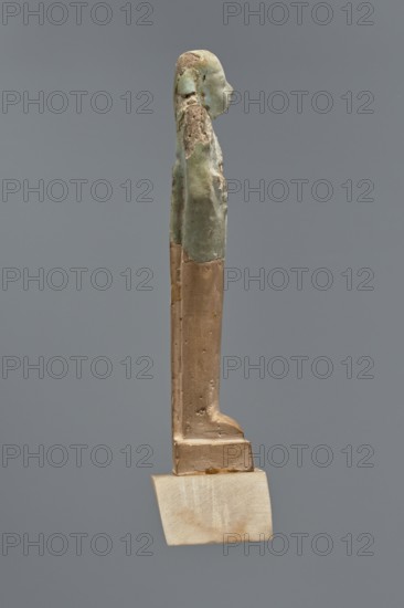 Fragment of a shabti, Mummy-shaped figurine, grave good, hoe held in hands. Upper part preserved to the beginning of the thighs. Smooth tripartite wig, back pillar at the back. Facial features carelessly rendered. Celadon glaze. Ptolemaic Period, 332–30 BC; El-Kantara. Shabti, height 7.8 cm., A slender vertical sculpture of a standing human figure shown in profile, mounted on a small rectangular base; the upper portion of the figure has a textured, weathered surface with greenish and pale blue tones mixed with areas of brown and beige, while the lower portion and base display smoother surfaces in warm tan and light brown shades; the background is a uniform mid-gray.
