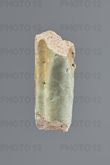 Fragment of a shabti, Middle part of the torso of a shabti figurine, preserved from the elbows to mid-thighs. Visible hands with hoe tips, traces of an engraved column of inscription on the front, back pillar on the back. Celadon glaze. 332–30 BC; Ptolemaic Period. El-Kantara place of discovery. Shabti, height 4.6 cm. PMID: unknown., A fragment of a cylindrical object with a rough, porous interior and a smoother outer surface, shown against a plain gray background; the outer surface is predominantly pale green with areas of light beige and faint yellowish tones, small dark speckles, and a narrow vertical streak of muted red along one side, while the exposed broken ends reveal a light tan, sandy texture.