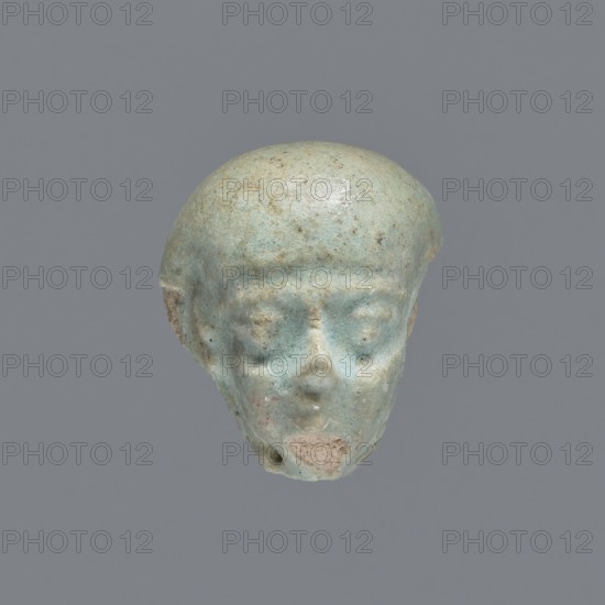 Fragment of a shabti; mummy-shaped funerary figurine. Head of a shabti figurine with carefully rendered facial features. Celadon glaze. Ptolemaic Period, 332–30 BC. From El-Kantara. Shabti; height 2.4 cm., A small sculpted head with a rounded top and simplified facial features, shown against a plain gray background; the object is primarily a pale bluish-green with areas of lighter and darker bluish tones and small patches of beige and brown, with a smoother sheen on the rounded scalp and a slightly rougher texture on the lower face, visible eye sockets, a subtle nose, and an eroded mouth area.