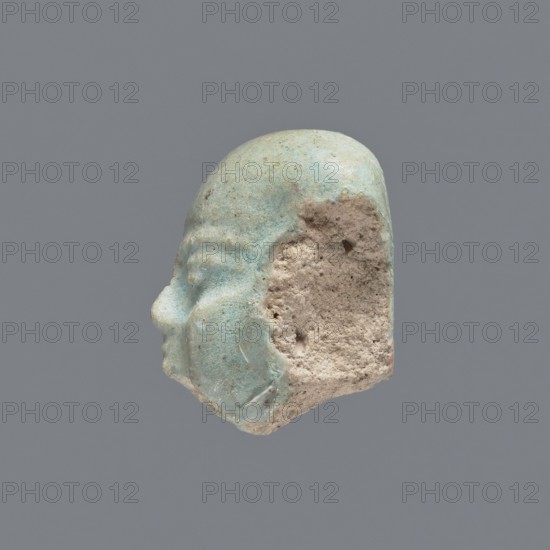 Fragment of a shabti;, Head of a shabti figurine with carefully rendered facial features. Celadon glaze. Mummy-shaped funerary figurine shabti. Ptolemaic Period, 332–30 BC. Found at El-Kantara. Height 2.4 cm., A small sculpted fragment of a human head shown in profile with visible facial features including a nose, closed eye, and rounded cheek, with a partially eroded rear section exposing a rough, porous interior; the outer surface is smooth and glazed in shades of pale blue and green with some areas of lighter cream and beige where the glaze is worn, while the exposed interior is a coarse, sandy beige; the background is a uniform gray.