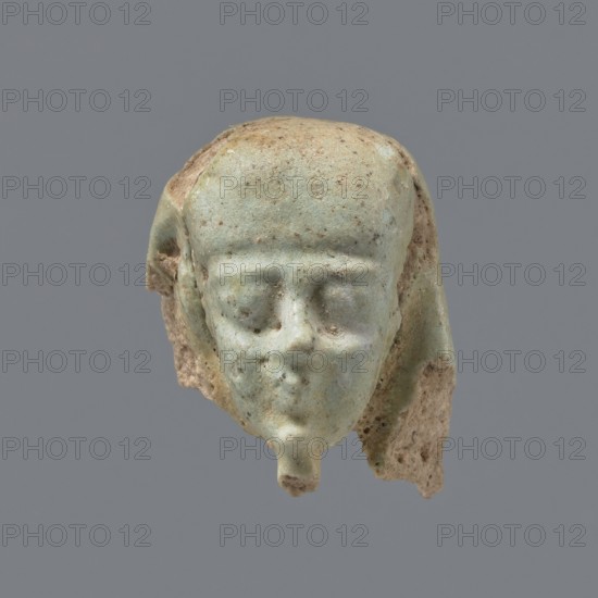 Fragment of a shabti. Head of a shabti figurine with carefully rendered facial features; smooth wig and thin beard. Celadon glaze. Ptolemaic Period, 332–30 BC. From El-Kantara. Shabti; height 2.9 cm., A small sculpted head fragment rendered in a pale greenish-blue with areas of beige and light brown, showing a rounded forehead, recessed eye areas, a defined nose, and a small protrusion at the chin; remnants of a head covering or headdress drape along the sides in similar greenish-blue and beige tones, and the surface shows pitting and weathered texture with speckles of brown against a plain gray background.