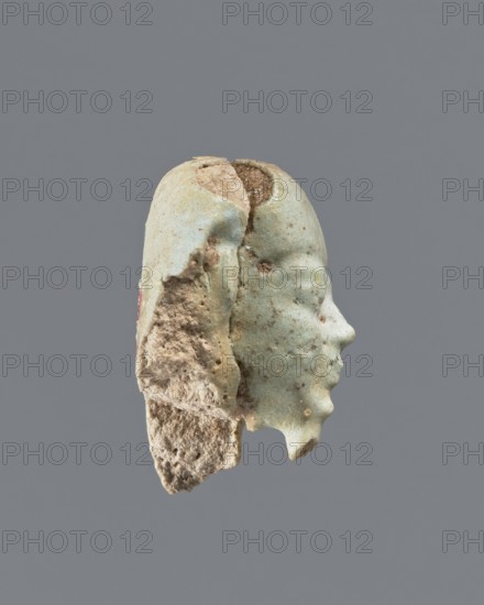 Fragment of a shabti. Head of a shabti figurine with carefully rendered facial features; smooth wig and thin beard. Celadon glaze. Ptolemaic Period, 332–30 BC. El-Kantara. Shabti, height 2.9 cm., A fragmentary sculpted head suspended against a solid gray background, showing a right-facing profile with a pronounced nose and chin; the surface is a weathered pale green with patches of beige and light brown where the material is eroded, revealing a rough, porous interior texture.