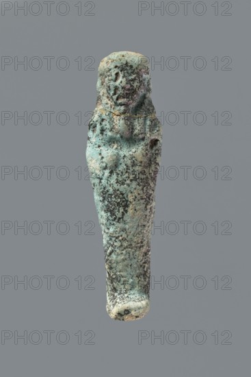 Ushabti, Mummy-shaped figurine, flat back. Arms crossed on the chest with only the hands protruding from under the shroud, holding hoes in both hands. Tripartite wig; facial details illegible. Not inscribed. 664–332 BC, Late Period, Memphis, Egypt. Height 8.5 cm., A weathered small statue shaped like a standing human figure with a rounded head and tapered body, showing rough pitted texture and areas of erosion; the surface displays a patina with shades of green and turquoise, darker green and black speckling, and hints of brown and beige; the figure’s facial features and limbs are worn but faintly discernible, and the object is set against a plain gray background.