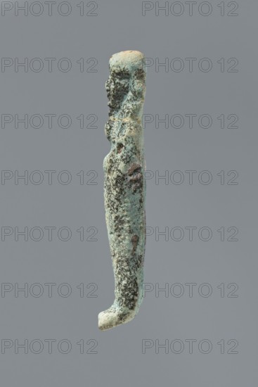 Ushabti, mummy-shaped figurine. Flat back. Arms crossed on the chest, only the hands protruding from under the shroud, holding hoes in both hands. Tripartite wig on the head. Facial details illegible. Not inscribed. 664–332 BC Late Period, Memphis, Egypt. Height 8.5 cm., A vertically oriented elongated object with an irregular, slightly curved shape that resembles a narrow figure; its surface is rough and pitted with textured areas and small raised bumps. The upper portion has a rounded top and a suggestion of shallow contours on one side; the middle narrows slightly and then widens into a gently rounded lower section that curves outward at the very end. The color palette is predominantly greenish-blue with areas of darker green and black mottling, interspersed with patches of pale beige and subtle brown tones. The background is a uniform mid-gray.