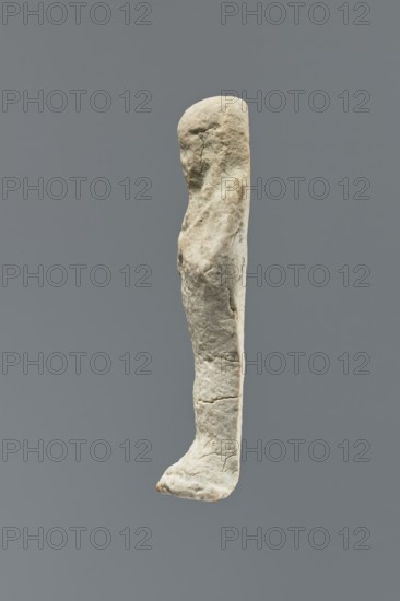 Ushabti, Mummy-shaped figurine standing on a base, flat back. Arms crossed on the chest, right folded over left; a pickaxe in the left hand and a hoe in the right. Smooth three-part wig, ears uncovered; facial details illegible; long beard damaged. No inscription. Date: 664–30 BC Late Period–Ptolemaic Period. Place of origin: Memphis, Egypt. Shabti; height 6 cm., A vertical, elongated sculptural object resembling a simplified human leg and foot is centered against a uniform gray background; the object has a rough, weathered surface with irregular textures and small cracks, and its color appears mainly off-white to pale beige with subtle darker beige and grayish patches and a small hint of muted brown at the tip of the foot.
