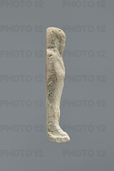 Ushabti, Mummy-shaped figurine, standing on a base with a flat back. Arms crossed on the chest, right folded over left; a pickaxe in the left hand and a hoe in the right. Wearing a smooth, three-part wig with ears uncovered. Facial details illegible; long beard damaged. No inscription. Date: 664–30 BC Late Period–Ptolemaic Period. Place of origin: Memphis, Egypt. Shabti, height 6 cm., A pale beige weathered vertical object with an irregular, elongated rounded top and a slightly protruding lower end resembling a curved foot, showing a rough, pitted surface with small cracks and textured indentations; a faint hint of muted red is visible along one side, and the background is a uniform medium gray.