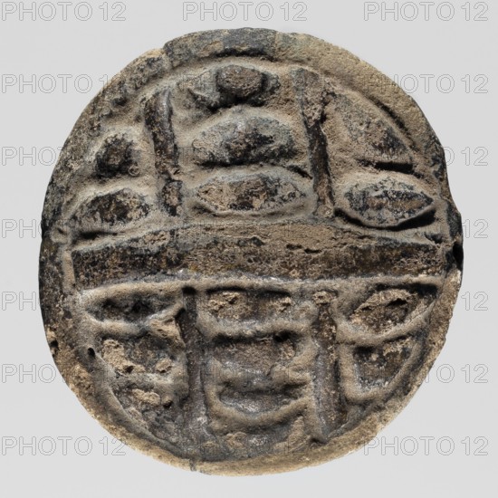 Amulet - multiple eye iret, eyes, protective amulet, ca. 818–715 BC; Third Intermediate Period, 23rd–25th Dynasty, Edfu, amulet, height 4.8 cm, width 4.5 cm, A round, flat object with a weathered, carved surface showing shallow raised and recessed shapes; the material appears rough and stone-like with a grainy texture. Visible colors include shades of gray, dark gray, beige, brown, and hints of black. The surface shows rounded and oval raised forms arranged in horizontal bands, separated by a raised horizontal ridge across the middle. A few vertical raised elements intersect the bands, and there are several irregular pits and erosion marks across the face. The outer edge is slightly irregular and worn, with areas of darker discoloration along the rim. The background is a uniform light gray.