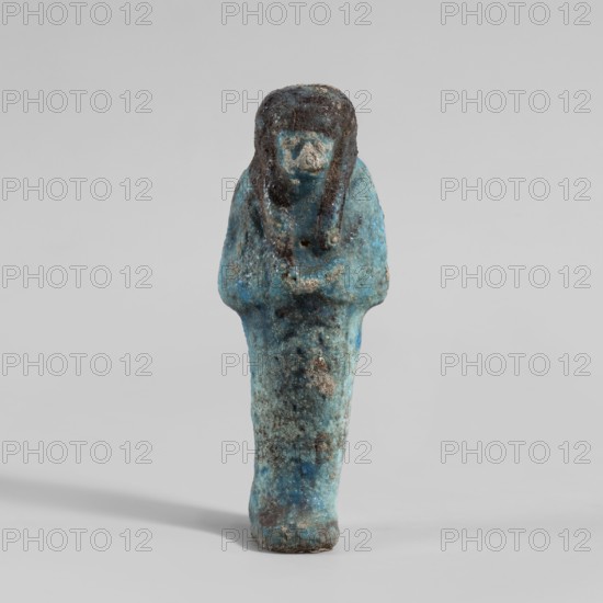 Ushabti, Mummy-shaped figurine. Hands folded on the chest—right over left. Hoes in each hand. Large basket slung over both shoulders on the back. Painted tools. Three-part smooth wig with a headband tied at the back; painted wig. Facial details blurred. Inscription engraved and filled with black dye on the front of the legs, now almost invisible. Third Intermediate Period, Deir el-Medina. Shabti. Height 10.7 cm, width 4 cm., A small statuette in shades of blue and turquoise with patches of darker blue and brown, shown upright on a light gray background; the figure has a rounded head with darker brown areas suggesting hair or a headdress, a simplified face with a lighter blue tone, narrow shoulders and arms folded across the chest, and a tapered lower body with mottled blue, turquoise, and brown surface texture and some rough, worn spots.