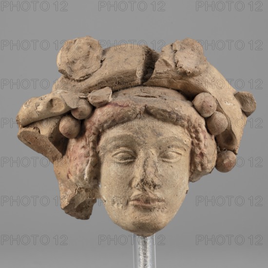 Head of Dionysus, unknown author. A small terracotta head of Dionysus, part of a larger lost sculpture. The figure has a large, full mouth, almond-shaped eyes with distinct eyelids, and malleable eyebrow arches; the nose is damaged. Losses are visible on the chin, above the left eyebrow, and on the right cheek. Hair, partially covered, frames the head with a central parting at the forehead. Head decoration of a ribbon and rosettes is partially preserved; identification though uncertain is suggested by the elaborate headdress. Traces of red-brown polychrome remain on areas such as the hair and lips. Dated 5th–4th century BC previously 3rd quarter of the 5th century BC. Place of origin: Tarentum. Figurine; height 6.8 cm, width 9 cm, depth 4.5 cm., The image shows a weathered sculpted head made of a tan-brown material mounted on a slender clear support, featuring a stylized face with closed eyes and a neutral mouth, and surrounded by a headdress or hair composed of rounded forms and drape...