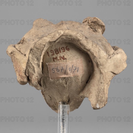 Head of Dionysus, A small terracotta head of Dionysus, part of a larger lost sculpture. The figure has a large, full mouth, almond-shaped eyes with distinct eyelids, and malleable eyebrow arches; the nose is damaged. Losses on the chin, above the left eyebrow, and on the right cheek. Hair, partially covered, frames the head with a central parting. Head decoration of a ribbon and rosettes, now partially preserved; identification though uncertain is suggested by the elaborate headdress. Traces of red-brown polychrome on hair and lips. Dionysus mythology, wreaths, Greek deities. Date: 5th–4th century BC formerly dated to 3rd quarter of the 5th century BC. Place of origin: Tarentum. Figurine. Dimensions: height 6.8 cm, width 9 cm, depth 4.5 cm., A worn, irregularly shaped object with a rough, claylike surface rests on a clear support against a neutral gray background; the object shows a concave central area surrounded by raised, lumpy edges and protrusions, with a small rectangular fabric ...
