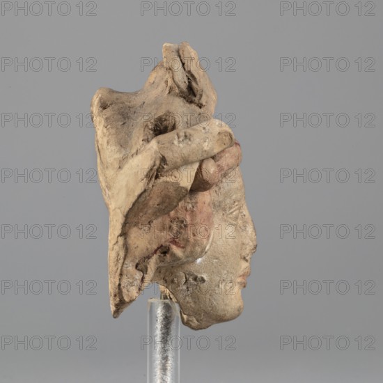 Head of Dionysus, A small terracotta head of Dionysus, part of a larger lost sculpture. The figure has a large, full mouth, almond-shaped eyes with distinct eyelids, and malleable eyebrow arches; the nose is damaged with losses on the chin, above the left eyebrow, and on the right cheek. Hair, partially covered, frames the head with a center parting. Head decoration consists of a ribbon and rosettes, now partially preserved; identification, though uncertain, is suggested by the elaborate headdress. Traces of red-brown polychrome are visible on fragments such as the hair and lips. 5th–4th century BC previously dated 3rd quarter of the 5th century BC. Tarentum. Terracotta figurine; h. 6.8 cm, w. 9 cm, d. 4.5 cm., A weathered sculpted fragment showing the profile of a human face mounted on a clear cylindrical stand, with a large irregular hat or headdress extending above and behind the head; the surface shows rough, eroded texture and traces of pigment in muted tones including beige, tan,...