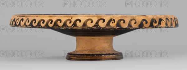 Red-figure plate depicting four fish on the interior, plate with an inwardly curled edge, Southern Italian pottery, plate vessel, red-figure technique, 2nd half of the 4th century BC, weight 0.488 kg., A shallow pedestal dish with a wide, flat rim decorated with a repeating wave-like pattern, showing wear and small chips; the dish is earthy tan with black painted decoration and black bands on the underside and base, and the surface shows variations of ochre and darker brown tones against a neutral gray background.