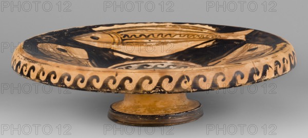 Red-figure plate depicting fish; plate with an inwardly curled edge. Interior painted with four fish. Southern Italian pottery, 2nd half of the 4th century BC previously dated 4th century BC. Southern Italy; plate vessel; weight 0.488 kg., A shallow round ceramic dish on a short pedestal, painted with a central fish motif and surrounding wave-like border; the dish shows areas of worn glaze and paint. The visible colors include beige, black, brown, and tan, with darker black used for the fish shapes and border patterns and lighter beige and tan for the background and pedestal, and some brownish aging marks.