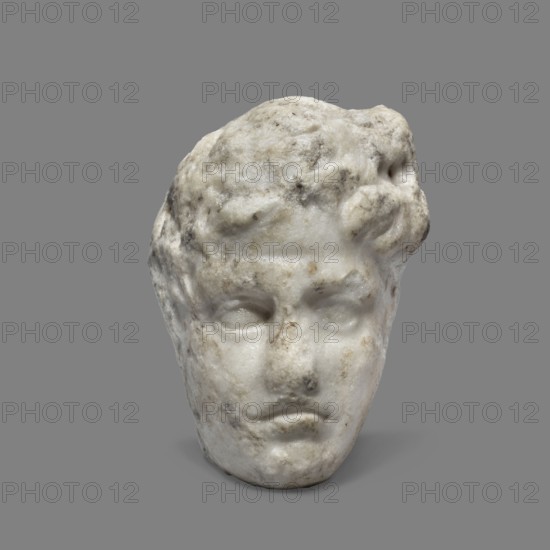 Head of a young man, unknown Greek workshop. A small head of a young man, probably a fragment of a bas-relief. The youth's face is oval, with almond-shaped eyes. A distinct furrow on the forehead. The mouth is twisted in a grimace. c. 350 BC, Greece. Sculpture; height 7 cm, width 6.5 cm, weight 0.51 kg., A worn marble head with textured surface and weathered features positioned against a flat gray background, showing a pale off-white and light gray stone with areas of darker gray and brownish discoloration, with sculpted hair forming a rough mass around the forehead and temples, faintly defined eyebrows and eye sockets, a rounded nose with irregular surface, and a closed mouth with a subtle downturn.