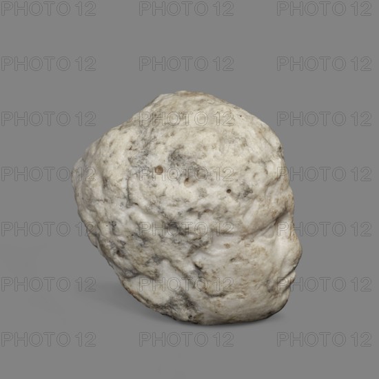 Head of a young man, unknown Greek workshop. A small head of a young man, probably a fragment of a bas-relief. The youth's face is oval, with almond-shaped eyes. A distinct furrow on the forehead. The mouth is twisted in a grimace. c. 350 BC, Greece. Sculpture; height 7 cm, width 6.5 cm, weight 0.51 kg., A single irregularly shaped object with a rough, pitted surface rests against a plain background; the object appears rounded with a subtle protrusion resembling a facial profile on one side. The surface shows a mix of pale white and off-white tones with areas of gray and light beige discoloration, and small darker gray spots and streaks scattered across it. The background is a uniform medium gray.