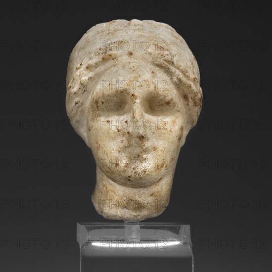 Head of a woman in a bonnet; Greek sculpture, female portrait, hairstyle; 5th century BC, Greece;, A close-up of a carved stone head displayed on a clear stand, showing a rounded face with hollowed eye sockets, a subtle nose and mouth, and a wrapped head covering; the surface is mottled with a mix of off-white, beige, tan, brown, and gray tones, and the background is dark gray.