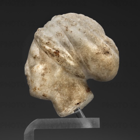 Head of a woman in a bonnet; Greek sculpture, female portrait; hairstyle; 5th century BC, Greece;, A carved stone head displayed on a clear stand against a dark background, showing the side profile of a face with a rounded bun at the back of the head; the stone surface is mottled with shades of white, beige, tan, and brown with areas of darker brown speckling and weathered texture, and the stand appears translucent with a metallic-looking support base.