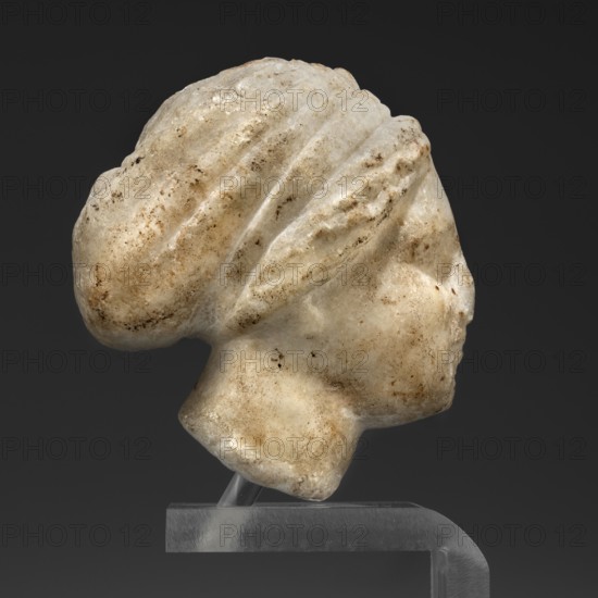 Head of a woman in a bonnet; unknown creator; Greek sculpture; female portrait; hairstyle; 5th century BC; Greece; Classified Gallic Scenography., A light-colored stone sculpture of a human head in profile displayed against a dark gray background, showing a rounded bun at the back with carved ridges suggesting hair, a smooth forehead and cheek, a rounded chin and neck, and surface discoloration and small dark brown and tan spots and stains on the stone; the sculpture rests on a clear support that sits on a darker translucent base, and visible colors include white, off-white, beige, tan, brown, clear, translucent gray, and dark gray.