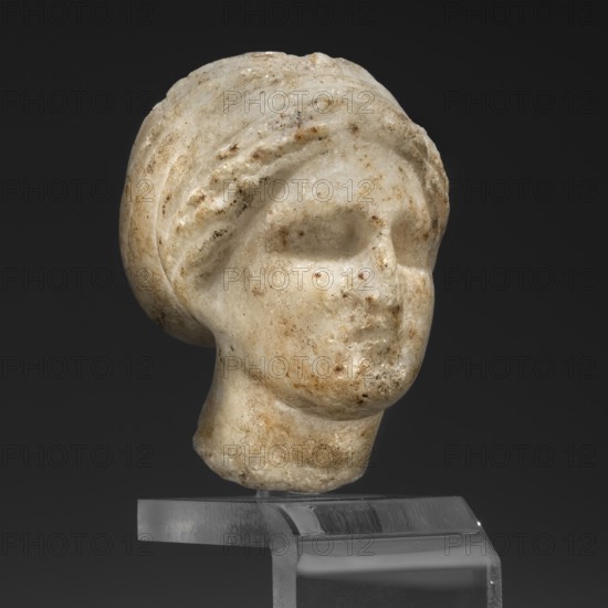 Head of a woman in a bonnet; Greek sculpture, female portrait; hairstyle; 5th century BC, Greece;, A carved stone head displayed on a clear stand against a dark background, showing a rounded hairstyle or head covering with layered ridges and a smooth neck; the surface is mottled with patches and speckles in beige, off-white, light brown, and darker brown tones, with worn and pitted areas on the cheeks, forehead, and chin; the facial features are softly modeled with shallow eye sockets and a subtle nose and mouth form.