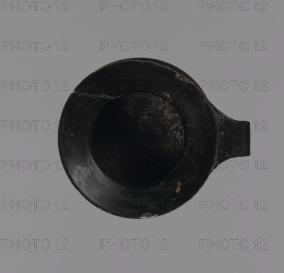 Gnathia-style mug, Antique vessel mug from Apulia, Southern Italy; 4th–3rd century BC. Red clay with glossy varnish, white-painted decoration and yellow retouching. Flat bottom, convex belly tapering to a wide spout; band-like rim. Decoration mainly on the upper belly opposite the rim: three elongated female heads in profile turned left with large eyes and hairstyles shaded with yellow, surrounded by an ornament of ivy fruit and leaves. Gnathia style; Greek/Ancient Greek pottery., A top-down view of a round vessel with a spout, the vessel appearing predominantly black with worn areas showing muted brown and beige tones, placed against a uniform gray background; the interior is dark with mottled lighter patches, and the rim shows a few small chips and scratches.