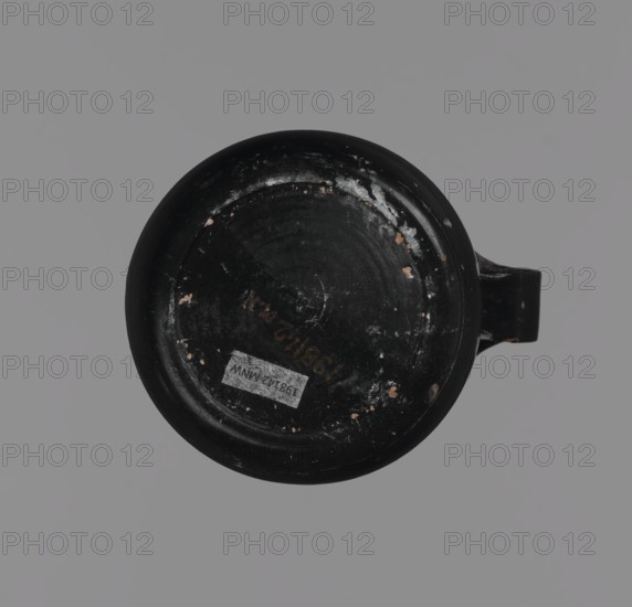 Gnathia-style mug, antique mug from Apulia Southern Italy, 4th–3rd century BC. Red clay with glossy varnish, white-painted decoration and yellow retouching. Flat bottom, convex belly tapering to a wide spout; band-like handle. Decoration mainly on upper belly opposite the handle: three elongated female heads in profile facing left with large eyes and hairstyles shaded with yellow, surrounded by an ornament of ivy fruit and leaves. Vessel cup., A round black object with a handle, seen from above against a gray background, showing worn and chipped areas with small spots of a light brown color and a few white scuff marks.