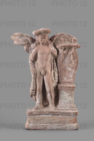 Figurine of Eros with a lyre, Eros myth., Greek deities, lyres musical instruments. Figurine of Eros standing on a base, holding a lyre resting on a base. 2nd century BC, Southern Italy . Terracotta figurine, height 19 cm, width 13.5 cm, depth 4.5 cm, weight 0.38 kg., A weathered terracotta figurine in shades of reddish-brown and pale pink stands against a flat grey background; the piece depicts a standing nude figure with outstretched wings and a draped cloth over one shoulder, a wreath or circular headpiece, and a raised arm touching the face, positioned beside a carved plinth or stele with ornamental relief, all resting on a rectangular stepped base showing mottled discoloration and surface wear.