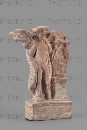 Figurine of Eros with a lyre, Eros myth., Greek deities, lyres musical instruments; Figurine of Eros standing on a base, holding a lyre resting on a base; 2nd century BC; Southern Italy ; material: figurine; height 19 cm, width 13.5 cm, depth 4.5 cm, weight 0.38 kg, A small weathered statue of a winged figure standing on a rectangular base, rendered in a warm terracotta tone with areas of lighter and darker wear and pale whitish residue; the figure appears nude with a cloak draped over one shoulder and one wing extended, showing rounded, worn features and smoothed facial details; beside the figure is a carved support or plinth with decorative rounded relief elements, sharing the same terracotta color and aged surface; the background is a flat neutral gray.