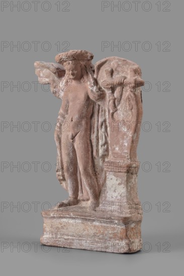 Figurine of Eros with a lyre, Eros myth, Greek deity, lyre musical instrument. Figurine of Eros standing on a base, holding a lyre resting on a base. 2nd century BC, Southern Italy, figurine, height 19 cm, width 13.5 cm, depth 4.5 cm, weight 0.38 kg., A small weathered statue of a winged figure standing on a rectangular base, shown against a plain gray background; the figure appears to have a rounded head and textured hair or headdress with wings extending from the shoulders, one wing partially visible to the left and the other forming a curved backdrop to the right, and the body and drapery show worn, uneven surfaces and soft contours; a vertical architectural element beside the figure has carved relief details and a rounded top, and the base shows layered tiers and rough texture. The visible colors are muted terracotta, light pinkish-brown, pale white weathering, and gray.