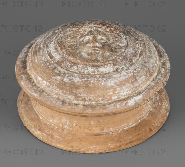 Pyxis lid, 2nd century BC, Southern Italy, vessel lid, weight 0.25 kg, height 6 cm, diameter 10.9 cm, A circular terracotta object with a stepped, rounded profile showing weathered surface texture and flaking. The top center features a raised, sculpted human face in low relief. The piece displays a mix of earthy tan and brown tones with patches of white encrustation and lighter beige wear. The background is a flat gray surface.