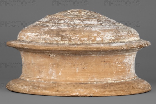 Pyxis lid, 2nd century BC, Southern Italy, vessel lid, weight 0.25 kg, height 6 cm, diameter 10.9 cm, A wide, rounded ceramic object with a domed top and a flared base sits against a plain background. The surface shows a weathered texture with irregular patches of lighter residue and areas of smoother wear. The piece displays earthy tones including tan, beige, and light brown, with spots of off-white and pale gray scuffing. The shape features concentric ridges on the dome and a pronounced rim midway, with subtle imperfections and small chips along the edges. The background appears uniform and gray.