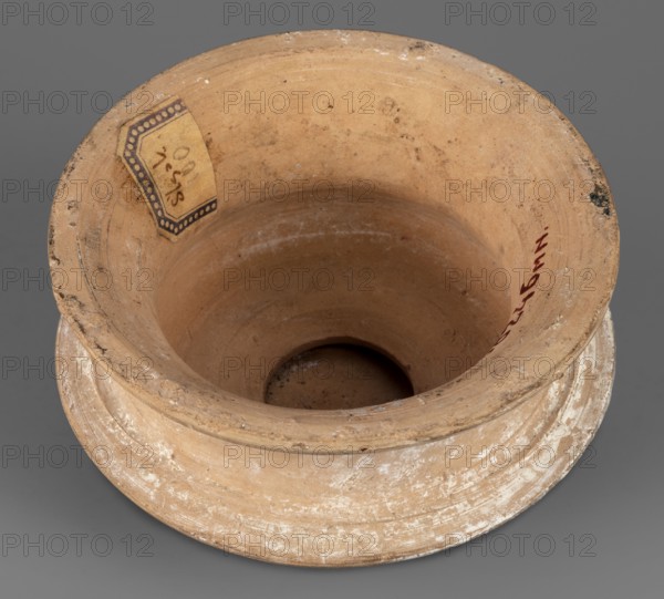 Pyxis lid, 2nd century BC, Southern Italy, vessel lid, weight 0.25 kg, height 6 cm, diameter 10.9 cm, A worn shallow bowl-shaped clay vessel viewed from above with an outward-flaring rim and a wide flat base, showing an interior depression leading to a small central hole; the clay surface is mostly light tan with areas of darker tan and beige discoloration and white spotting, and there is a small paper label adhered near the rim with a patterned edge; the rim shows rough, chipped areas and some blackened spots, and the vessel rests on a neutral gray background.