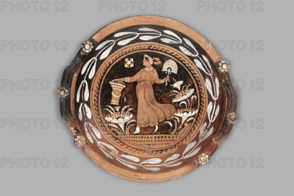Red-figure patera-phiale depicting a woman with a fan and basket. Classified Gallo-Classical Scenography, female images iconographic. Southern Italian pottery. Repossession Zabkowice Slaskie. Apulia Italy. Reddish clay with black glaze; numerous additions of white and yellow paint. Deep bowl with two vertical, profiled handles on the rim, decorated with three decorative knobs with heads adorned with white rosettes. Funnel-shaped foot with an upturned, profiled rim; similar profiling on the vessel rim. Richly decorated interior: in a medallion framed by laurel-branch motif painted in white and a black chasing-wave ornament, a woman running left in a chiton holding a basket and a fan. Stylized plants on the sides; a rosette in the left field. Dated 350–325 BC previously 3rd quarter of the 4th century BC. Type: vessel, patera. Weight: 1.144 kg., A round decorative plate with a central painted scene featuring a standing woman in a flowing dress holding an object in each hand, surrounded by...