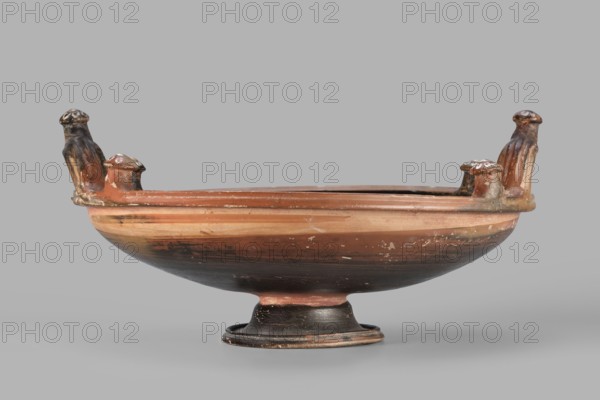 Red-figure patera-phiale depicting a woman with a fan and basket. Classified Gallo-Classical Scenography; female images iconographic. Southern Italian pottery from Apulia, 350–325 BC. Reddish clay with black varnish and numerous additions of white and yellow paint. A deep bowl with two vertical profiled handles on the rim, decorated with three decorative knobs topped by heads adorned with white rosettes. Funnel-shaped foot with upturned profiled rim; similar profiling on the vessel rim. Richly decorated interior: in a medallion framed by laurel-branch motif painted in white and an ornament of black chasing waves, a woman running left in a chiton carrying a basket and a fan. Flanking stylized plants and a rosette in the left field. Vessel type: patera; weight 1.144 kg., inscriptions/author unknown. Repossession Zabkowice Slaskie noted., A shallow ceramic bowl on a footed base with a wide rim, showing a group of small molded vertical handles or projections around the rim that have rounde...