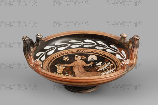 Red-figure patera-phiale depicting a woman with a fan and basket, Classified Gallo-Classical Scenography, female images iconographic. Southern Italian pottery; Apulia Italy. Reddish clay with black firnis; numerous additions of white and yellow paint. A deep bowl with two vertical, profiled handles on the rim, decorated with three decorative knobs with heads adorned with white rosettes. A funnel-shaped foot with an upturned, profiled rim; similar profiling on the rim of the vessel. Richly decorated interior: in a medallion framed with a motif of laurel branches painted in white and an ornament of black chasing waves, a woman running to the left in a chiton with a basket and a fan. On the sides are stylized plants; in the left field a rosette. Dated 350–325 BC previously 3rd quarter of the 4th century BC. Vessel type: patera. Weight 1.144 kg., A shallow ceramic bowl with a rounded foot and a wide rim, painted mainly in black and terracotta with white decorative elements, features a cent...