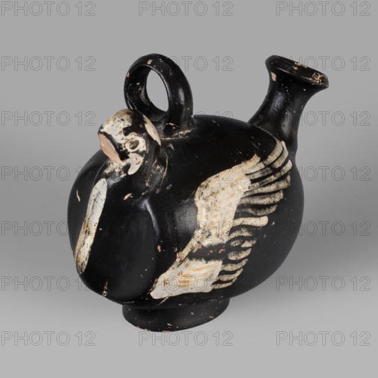 Guttus gnathia in the shape of a bird, bird with wings iconography, southern Italic pottery, end of 4th–beginning of 3rd century BC formerly ca. 300 BC, Apulia, guttus vessel; height 9.5 cm, width 6.4 cm, depth 11.3 cm, weight 0.164 kg., A rounded ceramic vessel with a short pedestal base and a small flared spout, finished predominantly in black with worn areas revealing beige and off-white patches, has a loop handle at the top and a sculpted bird-like head projecting from the shoulder; the bird motif features a beak area in a lighter beige and a wing rendered in layered off-white and beige strokes against the black body, with visible scuffs and chipped spots showing underlying terracotta and lighter surfaces, set against a neutral gray background.