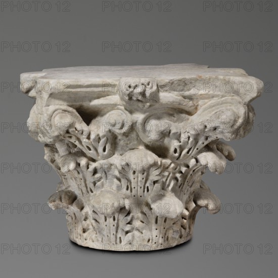 Corinthian capital, Sacred/Roman architecture; architectural element and decorative capital, 2nd century, Roman Empire 27 BC–476. Corinthian similar to kalathos capital with three rows of acanthus leaves; all leaves similar, wavy surfaces, divided by deep, softly bending grooves veins to the sides, rounded with lobed edges. Upper edge of the kalathos visible above the leaves. Abacus with extended corners composed of two plates of almost equal height. Round hole on the upper surface probably trace of contemporary mounting and a similar hole in the base. Great softness of modeling of details; flakes separated by holes. Rosettes present. Weight 133.5 kg., A carved stone capital with a weathered surface showing a group of acanthus-like leaves and spiraled volutes; the piece is primarily off-white and light gray with subtle beige and darker gray discolorations, and it sits against a smooth medium-gray background.