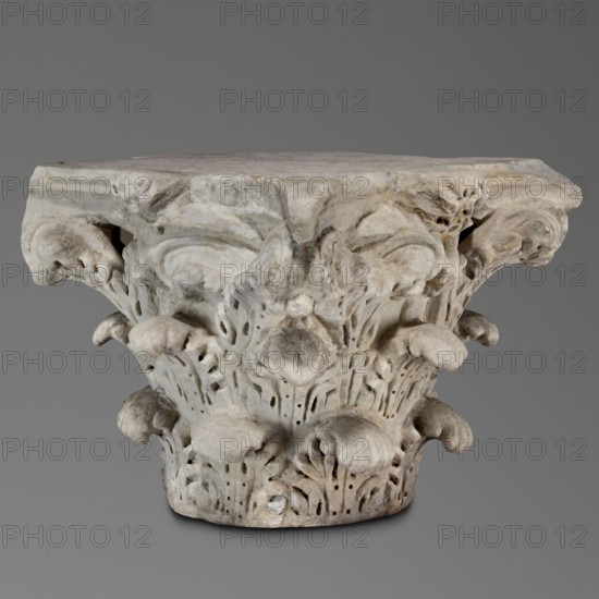 Corinthian capital, Sacred and Roman architecture; Corinthian capital similar to kalathos capitals. 2nd century, Roman Empire 27 BC–476., Materials and form: 3 rows of acanthus leaves, all similar with wavy surfaces, divided by deep, softly bending grooves veins to the sides; leaves rounded with edges divided into lobes. The upper edge of the kalathos is visible above the leaves. An abacus with extended corners composed of two plates of almost equal height. A round hole on the upper surface of the capital—probably a trace of contemporary mounting; a similar hole in the base. Great softness of modeling of details; flakes separated by holes. Weighs 133.5 kg. Tags/ornament: acanthus, rosettes., A carved stone column capital with ornate acanthus-like foliage and volute shapes sits against a plain gray background; the stone shows a weathered surface with a mix of light gray, off-white, beige, and subtle brownish tones, and the carving features rounded leaf forms, curled tendrils, perforated...