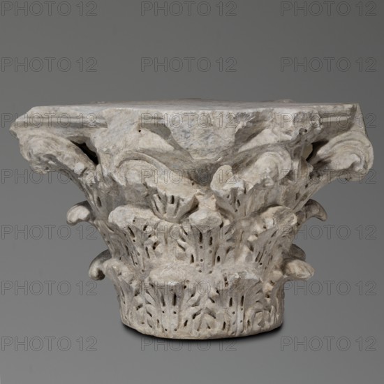 Corinthian capital, Sacred architecture, Roman architecture; capitals; acanthus ornament; rosettes. Corinthian capital similar to kalathos capitals. 3 rows of leaves. All leaves similar, with a wavy surface, divided by deep, softly bending grooves—veins—to the sides, rounded, with edges divided into lobes. The upper edge of the kalathos is visible above the leaves. An abacus with extended corners is composed of two plates of almost equal height. A round hole on the upper surface of the capital—probably a trace of contemporary mounting of the capital. A similar hole in the base. Great softness of modeling of details. Flakes separated by holes. 2nd century, Roman Empire 27 BC–476. Architectural element; element of architectural decoration; capital; architectural detail. Weight 133.5 kg., A carved stone architectural capital with a weathered surface, showing a group of ornate acanthus-like leaves and scrolls arranged around its cylindrical base and flaring top, with visible chips and roug...