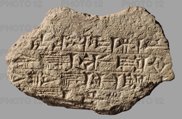 Fragment of a brick with a cuneiform inscription, architectural element, cuneiform writing; fragment with 4 lines impressed with a stamp of the inscription of the Neo-Babylonian king Nebuchadnezzar II Nabu-kudurri-usur, reign 604–562 BC Neo-Babylonian period. Archaizing graphic style of cuneiform characters. Findspot: Al Hillah ancient Babylon in Babylonia; possible Borsippa. Material: brick. Weight 470 g; max length 14.5 cm, width 9.1 cm, thickness 5.4 cm., A roughly oval, irregularly shaped stone slab with a textured, weathered surface in shades of beige, tan, and light brown sits against a flat gray background; the slab shows carved linear and wedge-shaped indentations across its face forming repeated horizontal groupings, with chisel marks and surface abrasions visible, and areas of shadow creating darker brown contrast within the carved grooves while the higher surfaces appear lighter beige.