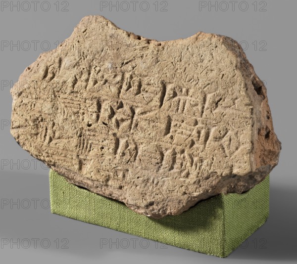 Fragment of a brick with a cuneiform inscription, architectural element, cuneiform writing, Near Eastern architecture, Eastern Gallic Scenography. Fragment of a brick with 4 lines, impressed with a stamp, bearing the inscription of the Neo-Babylonian king Nebuchadnezzar II Nabu-kudurri-usur, reign 604–562 BC. Archaized graphic style of cuneiform characters. Date: 604–562 BC reign of Nebuchadnezzar II, Neo-Babylonian period. Provenance: Al Hillah ancient Babylon, Babylonia; possibly Borsippa. Material: brick. Weight: 470 g. Dimensions: max length 14.5 cm; width 9.1 cm; thickness 5.4 cm., A rough, irregular stone fragment with a sandy beige surface rests on a textured rectangular block covered in green fabric; the stone shows a weathered, pitted texture with many shallow carved or incised marks and grooves across its face, and its edges are jagged and uneven; the background is a smooth gray, and the visible colors in the image include beige, light brown, green, gray, and small areas of d...