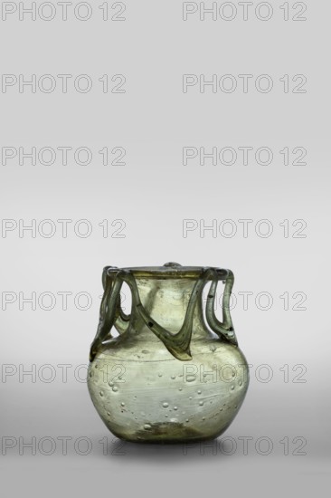Flask, unknown eastern workshop, glass vessel, miniature vessel, Type: Barag type VI:9. Transparent glass, yellowish-green with applied thread decoration between the arms and the rim edge in the form of 10 sharp bends; free-blown. Outer diameter of the rim: 5.50 cm. Bottom diameter: 3.90 cm. 4th century, Roman Empire 27 BC–476. Vessel, weight 0.076 kg., A small translucent vessel sits centered against a light gray background, the glass showing a greenish tint with areas of pale olive and subtle yellowish highlights; the body is rounded and bulbous with a narrower neck and a slightly flared rim, and a group of decorative looped handles of the same greenish glass drape from the rim down toward the shoulder creating flowing, curved shapes; the surface of the vessel contains numerous small bubbles and streaks, with some darker inclusions and faint horizontal striations, and the base appears slightly thicker and darker than the upper areas.