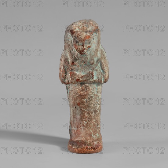 Ushabti, mummy-shaped figurine. Made of clay, painted cream with black details. Arms crossed on the chest, right hand folded over left; each hand holds raised hoes modeled in relief and painted black. Smooth tripartite wig with a black-painted headband; facial details marked in black. Rectangular basket on the back slung over both shoulders. Inscription on the front of the legs in one column, illegible. III Intermediate Period, 21st Dynasty, Deir el-Medina, shabti; height 11 cm, width 4.2 cm., A small standing figurine with a rounded head and simplified facial features, a blocky torso with short arms folded across it, and a tapered skirt-like lower body, showing a rough, pitted surface texture and irregular edges; the object displays a mix of greenish-blue patina, brownish rust tones, and areas of muted beige and gray, casting a soft shadow on a plain light gray background.