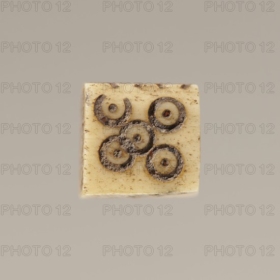 Game die made of animal bone with six unequal sides. Each side is engraved with a number from 1 to 6 in the form of circles with a dot in the center. 4th century, Egypt. Height 0.7 cm, width 0.7 cm, depth 0.7 cm., A small square object with an aged, creamy ivory surface showing a cluster of circular impressions in dark brown and black tones, some filled with a sandy, textured residue; the edges of the square show wear and slight discoloration in darker brown, and the background is a smooth, muted taupe-gray.