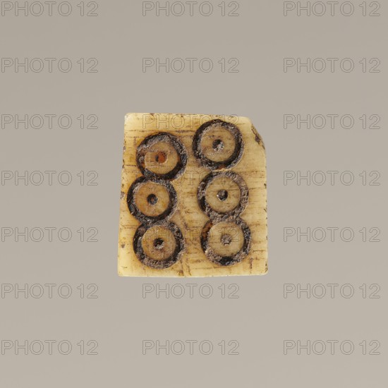 Game die made of animal bone with six unequal sides. Each side is engraved with a number from 1 to 6 as circles with a dot in the center. 4th century, Egypt. Height 0.7 cm, width 0.7 cm, depth 0.7 cm., A small rectangular object with rounded corners in shades of beige and tan sits against a plain light gray background; the object's surface shows a few darker brown circular impressions arranged in two vertical columns, each impression having a darker outer ring and a lighter inner circle, with some rough textured dark brown material within and around the rings, and faint horizontal striations visible on the beige surface.