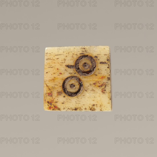 Game die made of animal bone with six unequal sides. Each side is engraved with a number from 1 to 6 as circles with a dot in the center. 4th century, Egypt. Height 0.7 cm, width 0.7 cm, depth 0.7 cm., A square, flat object with a worn, textured surface in creamy beige and yellow tones sits against a plain light gray background; the object shows several brown and dark brown circular markings with rough, crumbly edges and a few small areas of reddish and orange discoloration, along with many fine brownish streaks and speckles across its surface.